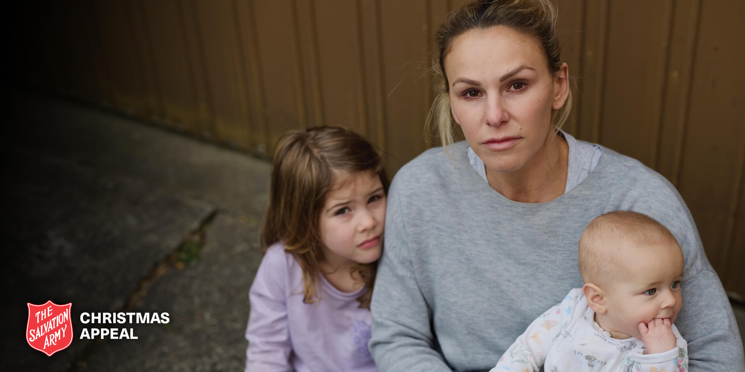 Amy with her daughter beside her and newborn in her arm. They are sitting on the sidewalk looking distressed.