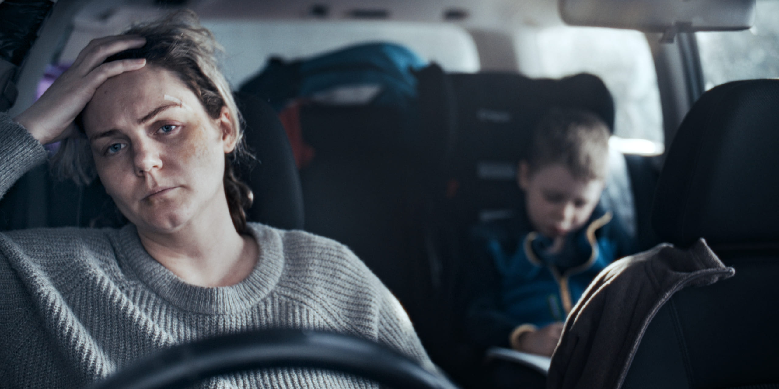 Mother in a car feeling stressed, with her young son sitting in the backseat.